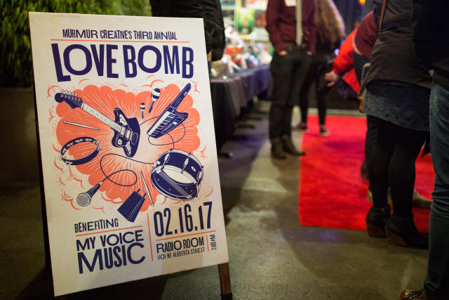 Murmur Creative's Love Bomb event sign featuring musical instruments, date, and beneficiary My Voice Music, set in a festive atmosphere.